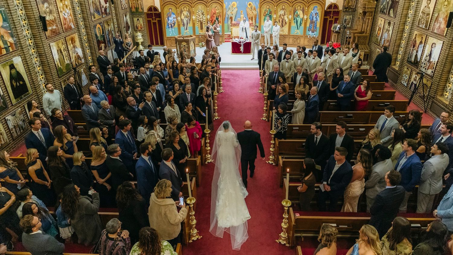 Bride walks down the aisle with escort in ornate church filled with wedding guests during ceremony.