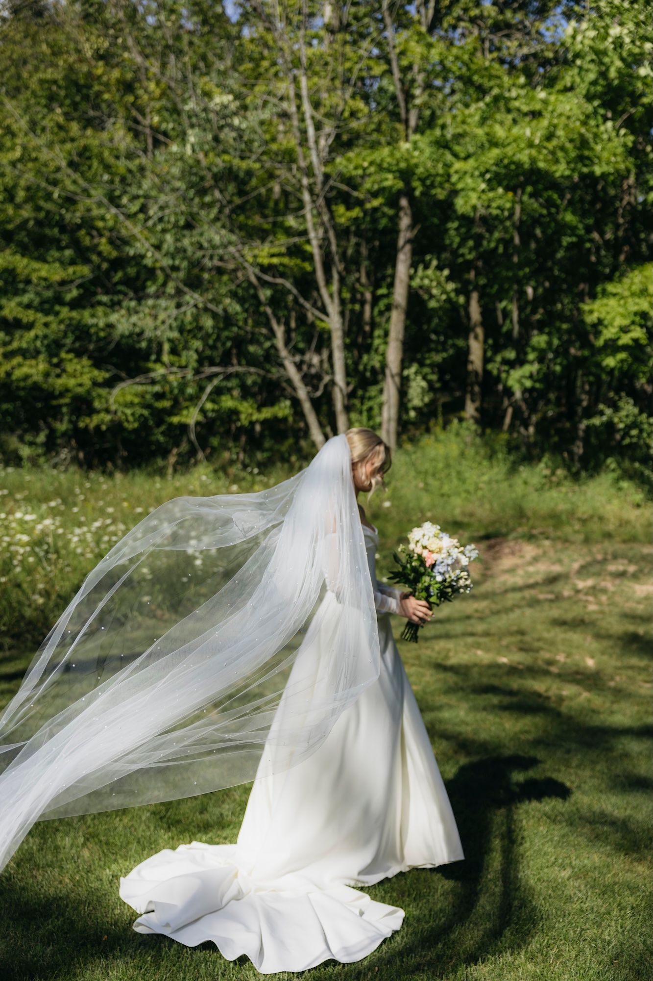 Bride in white gown and flowing veil holds bouquet outdoors on green lawn with lush trees in background.