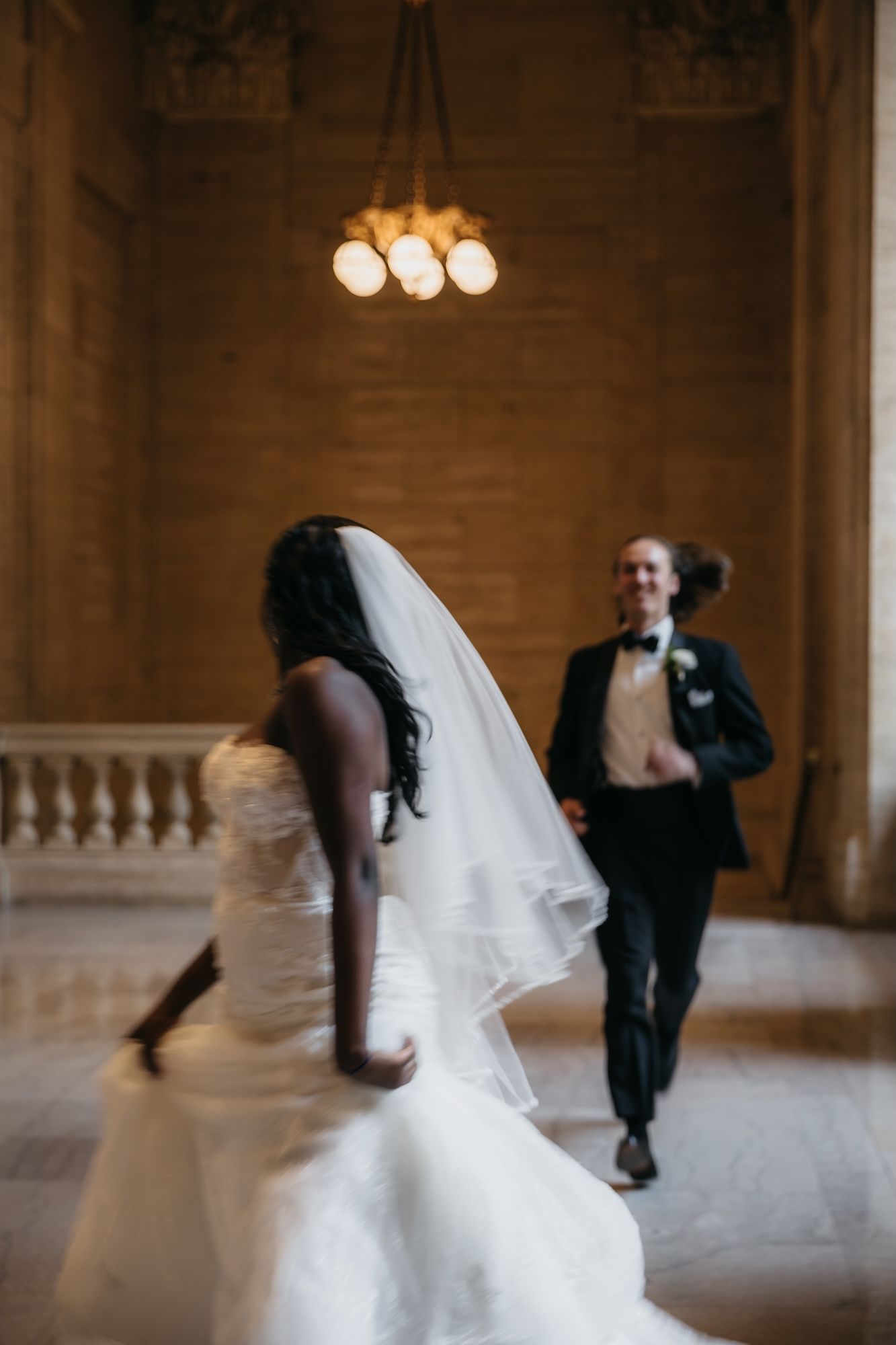 Bride in white gown and veil walks toward smiling groom in tuxedo inside elegant wedding venue with chandelier.