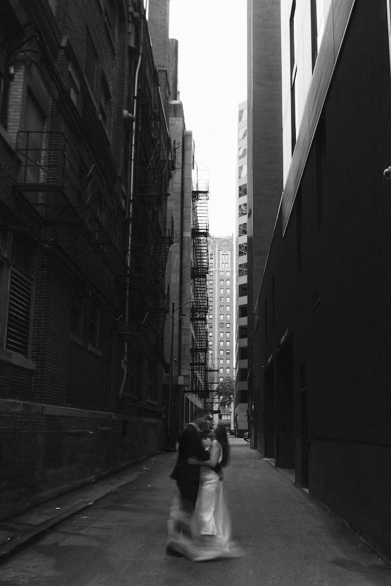 Black and white photo of a couple walking through a narrow urban alley with fire escapes and tall buildings.