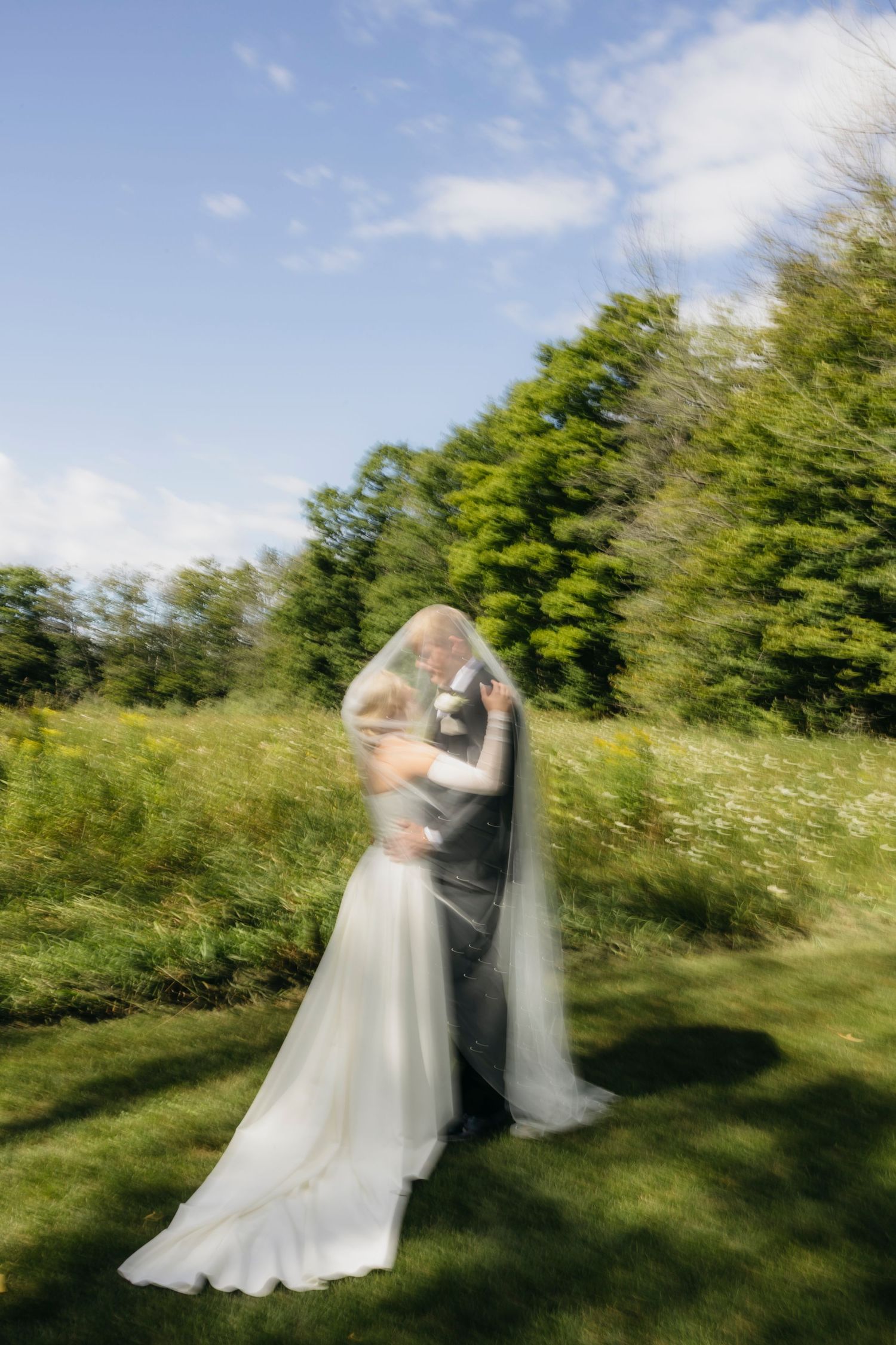 Bride and groom sharing a romantic kiss under a flowing veil outdoors on a sunny day with lush green trees.