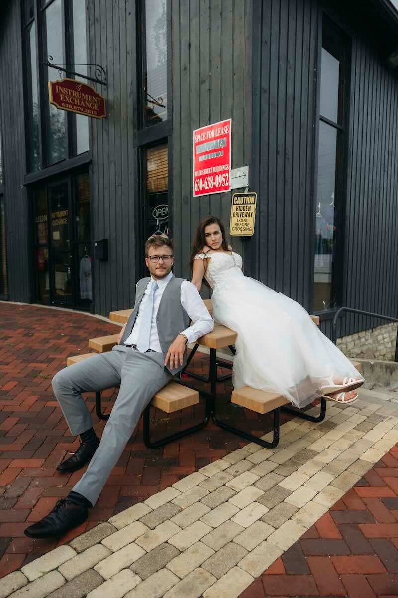 Bride in a white wedding gown and groom in a gray vest and suit lounging on a picnic table outside a dark building on a brick patio.