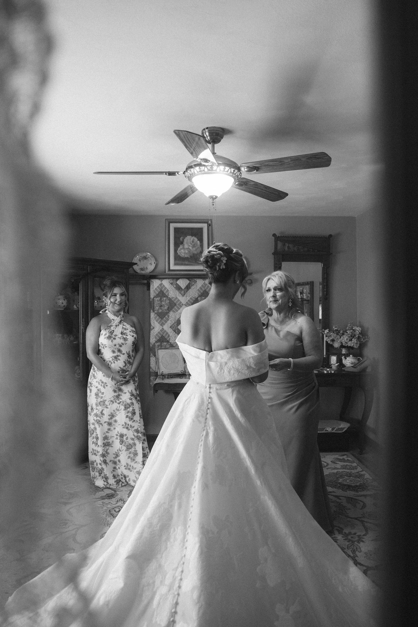 Black and white photo of a bride in a wedding gown with two women in a decorated room with a ceiling fan.