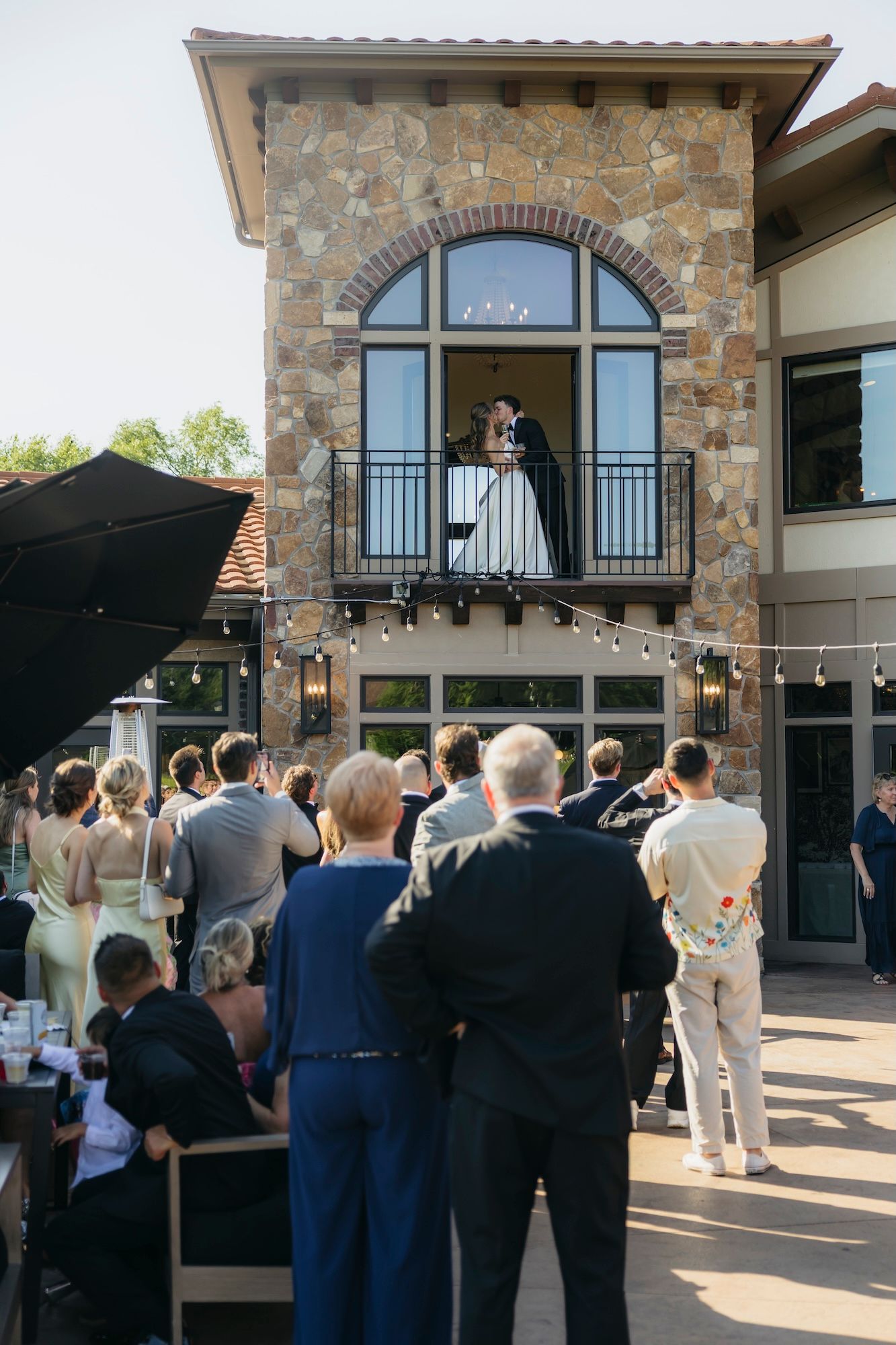 Wedding guests gather outdoors watching a couple on a stone building balcony during a romantic ceremony celebration.