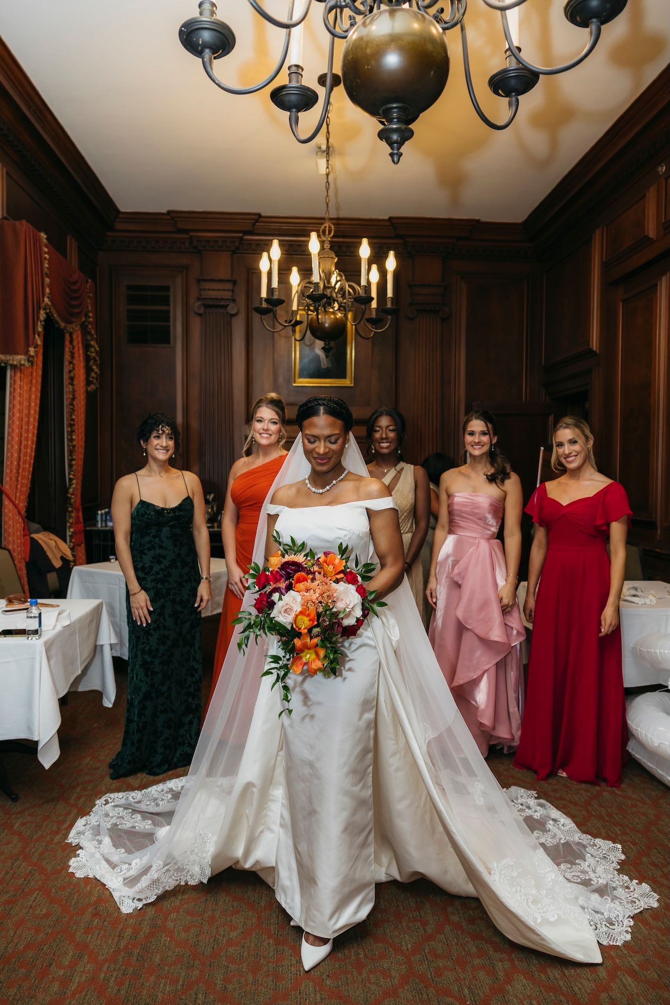 Bride in white gown holds colorful bouquet with bridesmaids in colorful dresses standing behind her in elegant wood-paneled room.