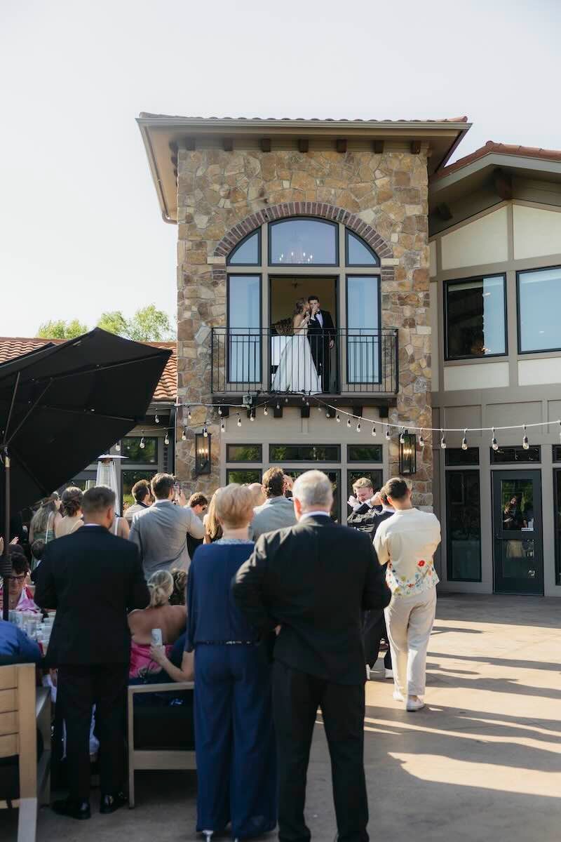 Wedding guests watch a bride and groom share a moment on a stone building balcony at an outdoor venue with string lights.