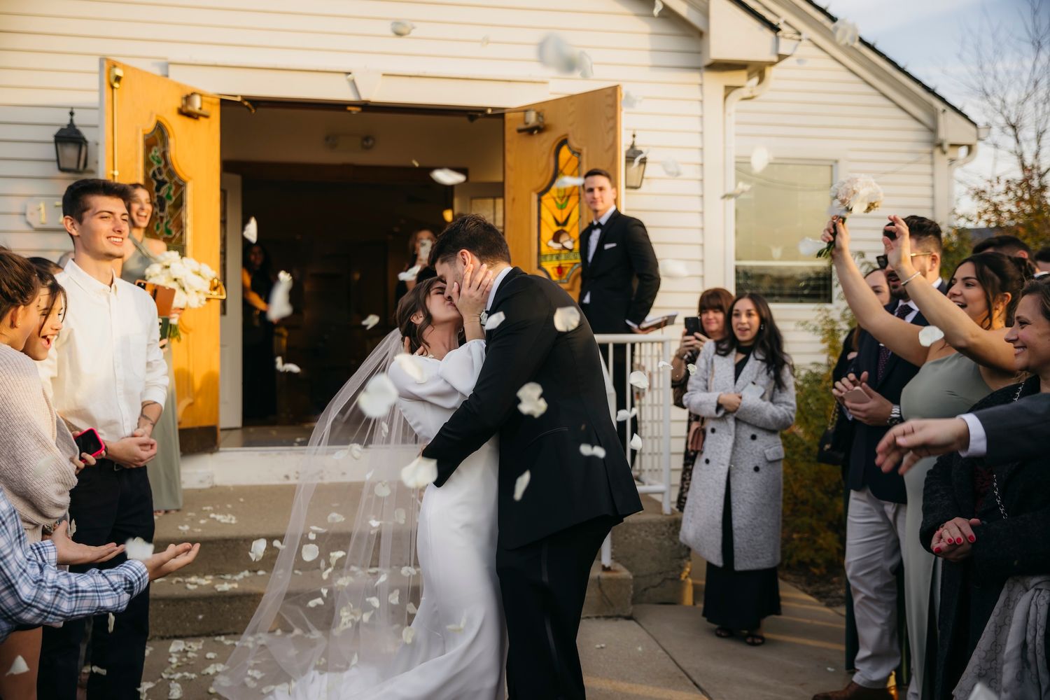 Bride and groom share a kiss outside a white chapel as guests toss flower petals during their wedding ceremony exit.