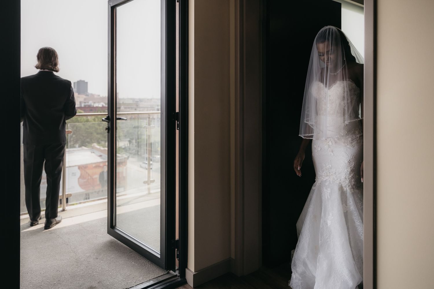 Bride in white mermaid gown and veil stands in doorway as groom looks out window on wedding day.
