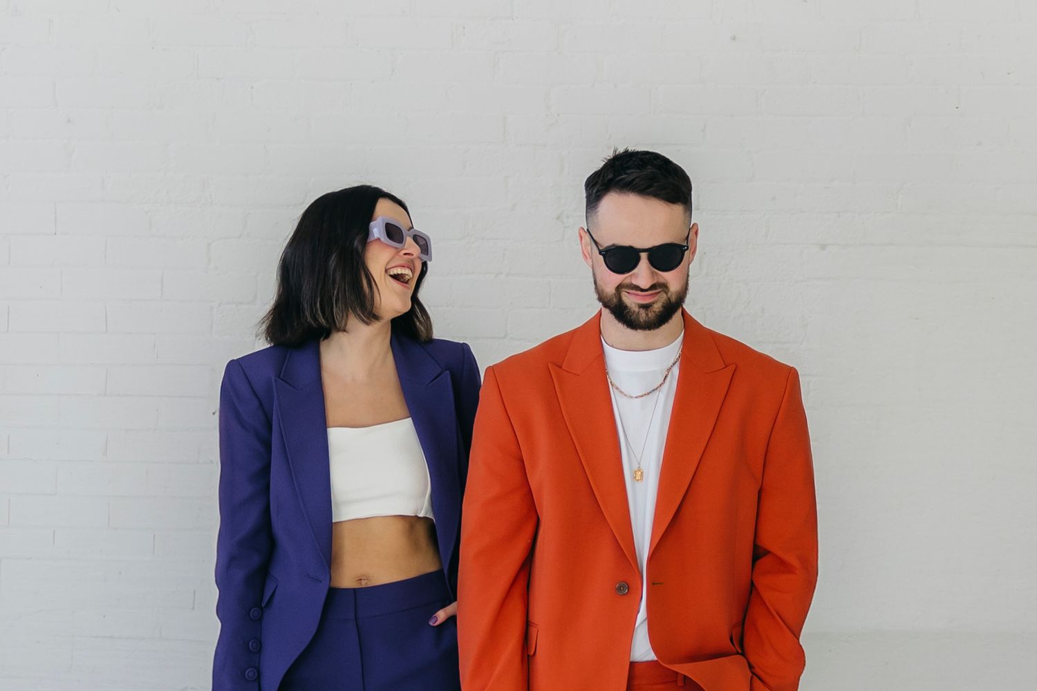 A woman in a purple blazer and a man in an orange blazer wearing sunglasses pose against a white wall.
