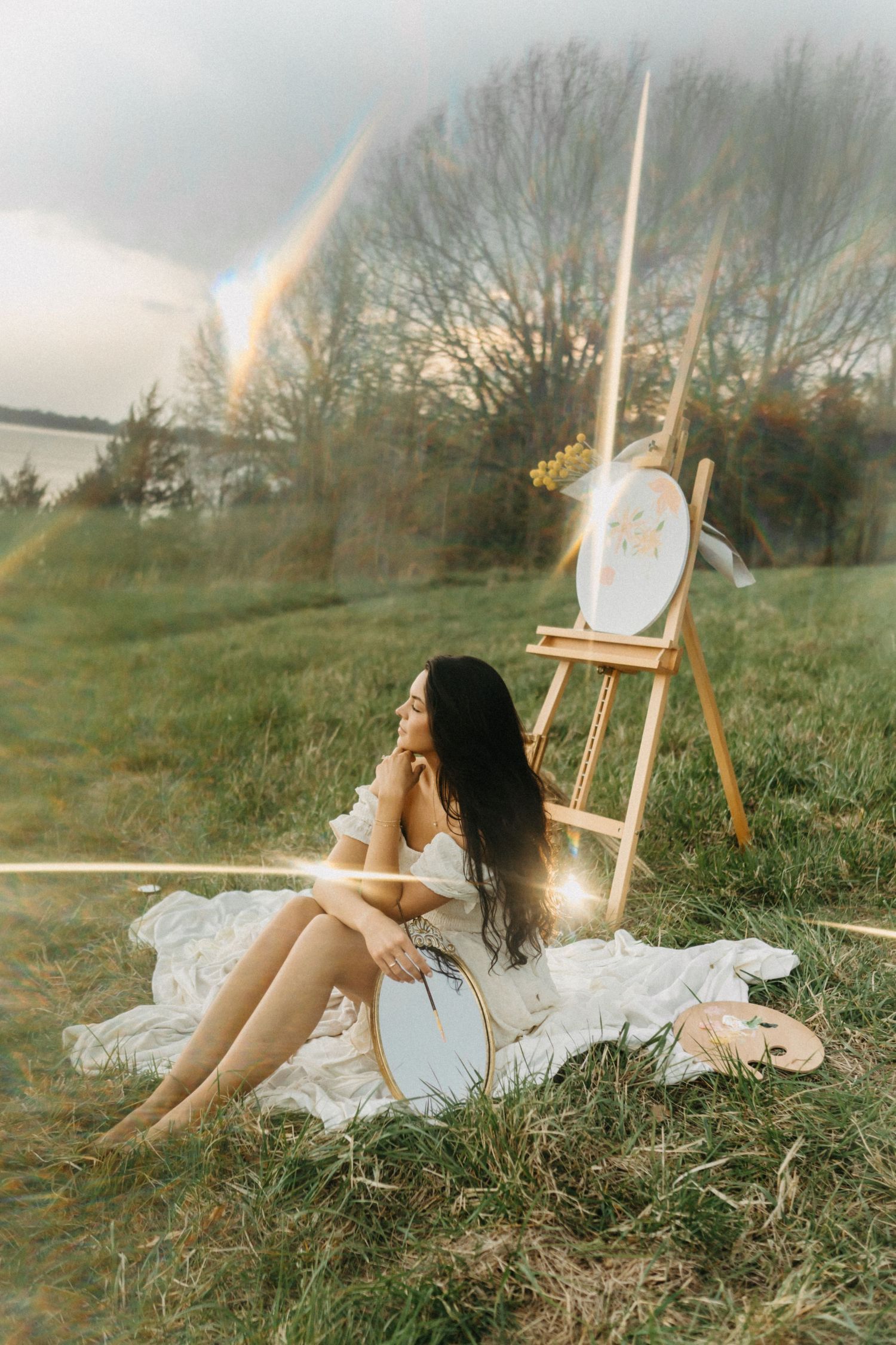 Woman in white dress sitting beside a wooden easel on grass with a rainbow backdrop.