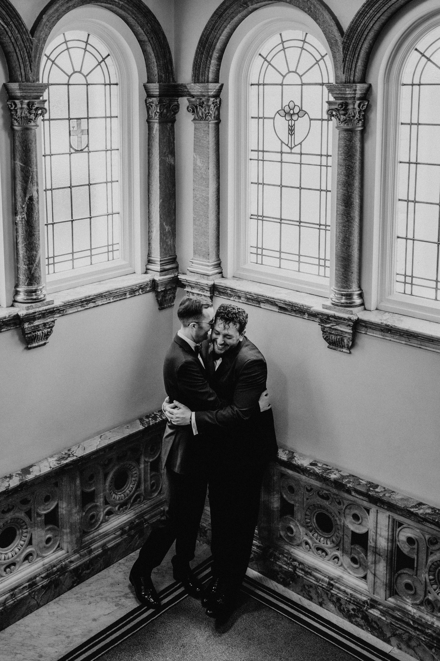 two grooms hug together on the staircase of Kimpton Fitzroy at their black tie London wedding