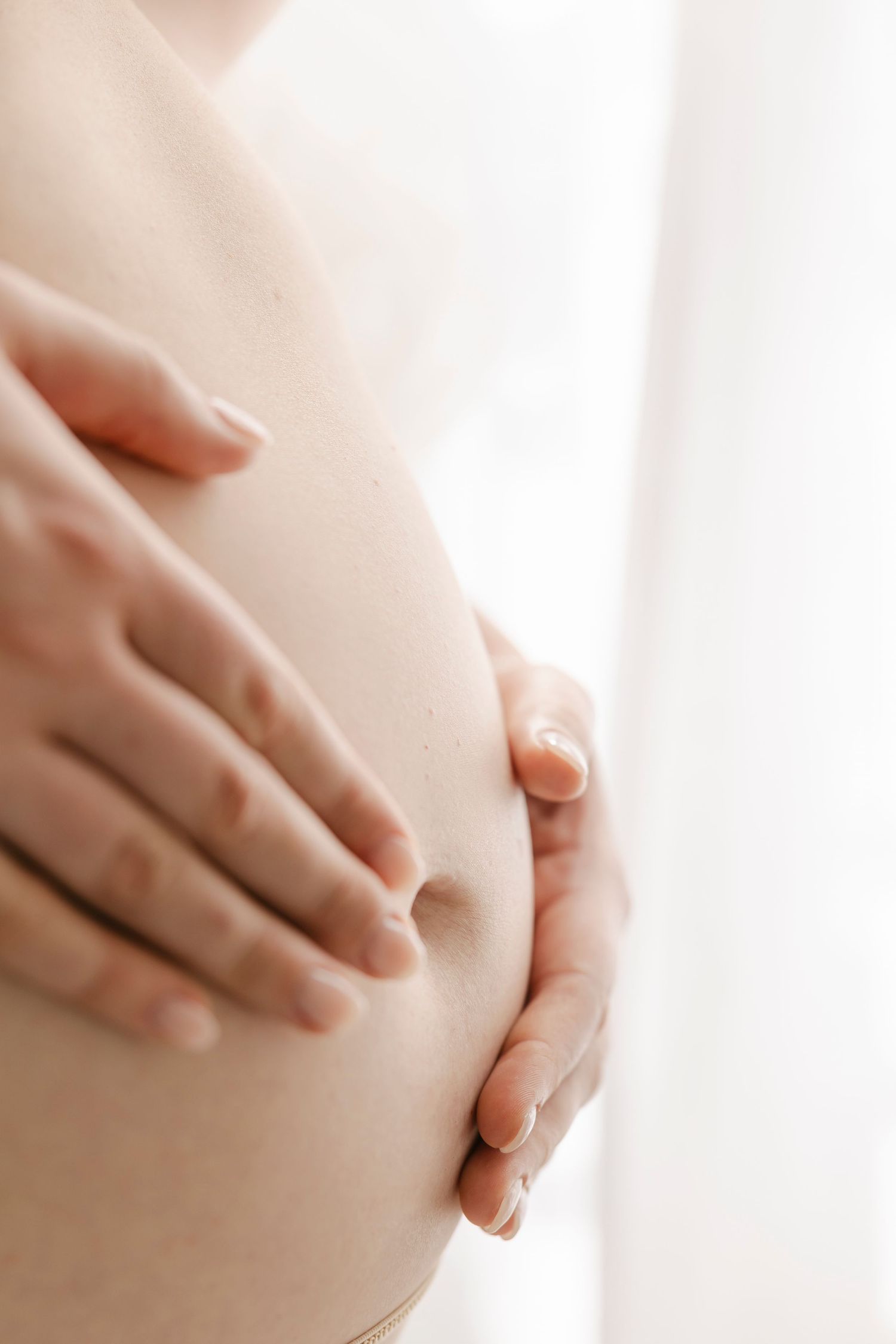 Close-up of a pregnant woman cradling her bare baby bump with both hands against a soft white background.
