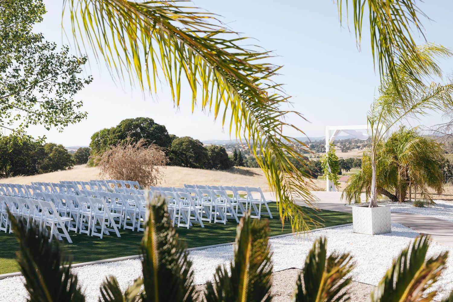 White wedding chairs arranged outdoors with palm trees and mountain views in the background.