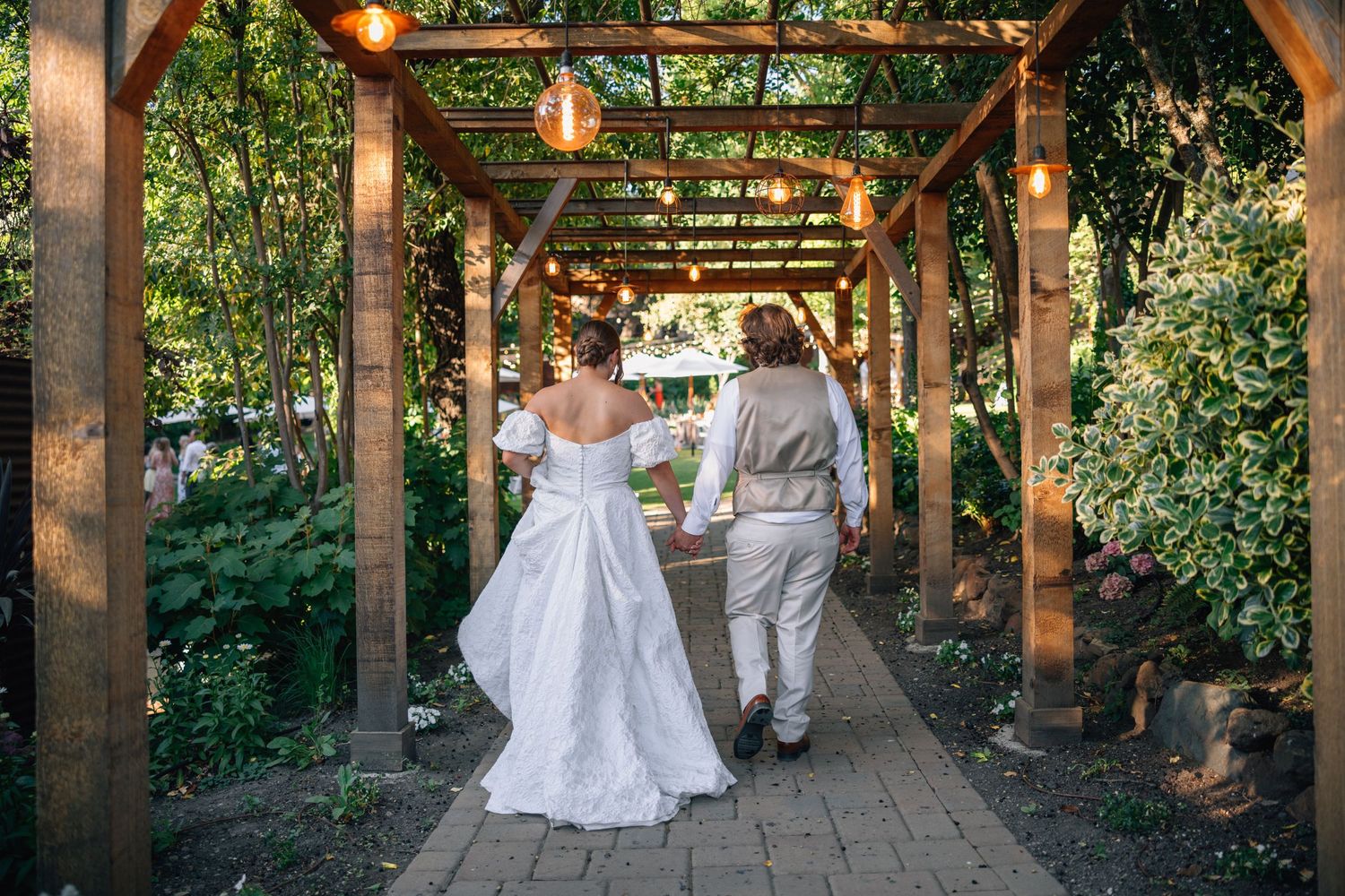Wedding couple walking under wooden pergola decorated with string lights and greenery.