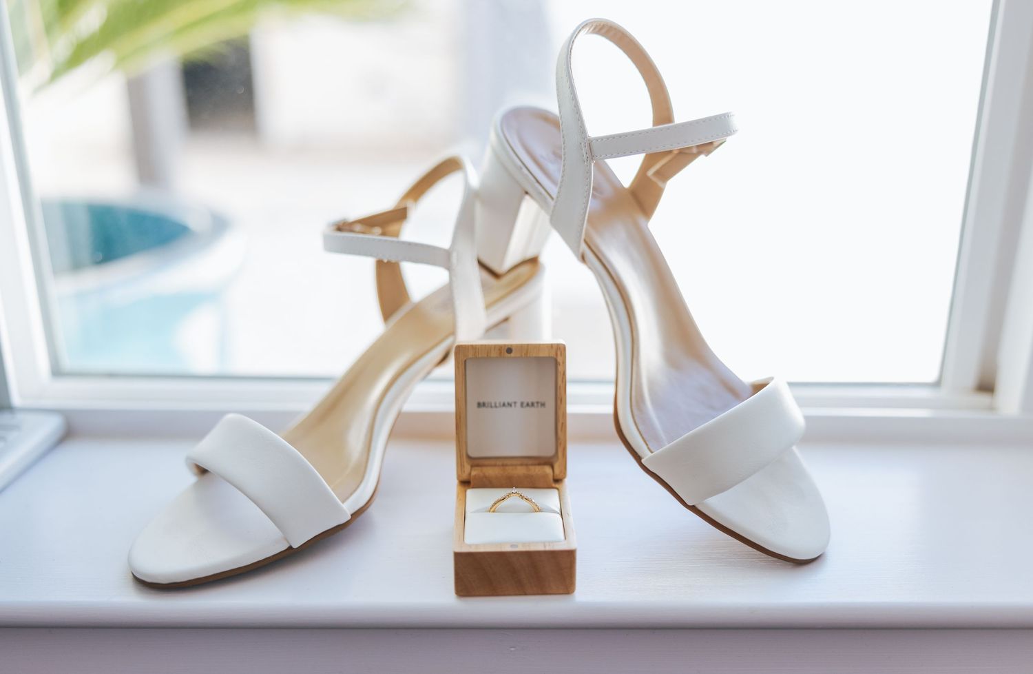 White bridal shoes and ring box displayed on windowsill in natural light.