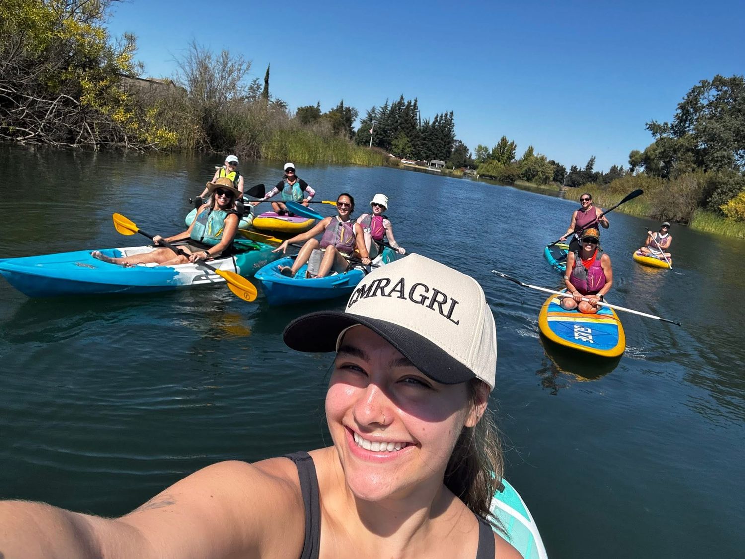 Woman in MRAGRL cap takes selfie while kayaking with group on calm river on sunny day.