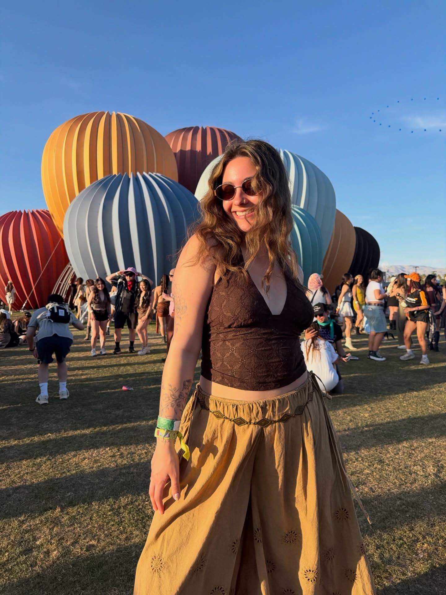 Woman in lace top and gold skirt smiles at outdoor festival with colorful hot air balloons in background.