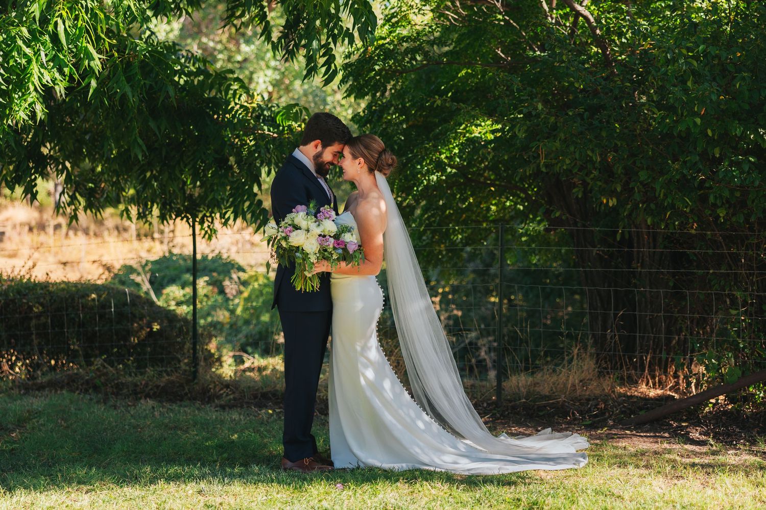 Wedding couple posing under green trees in garden setting.
