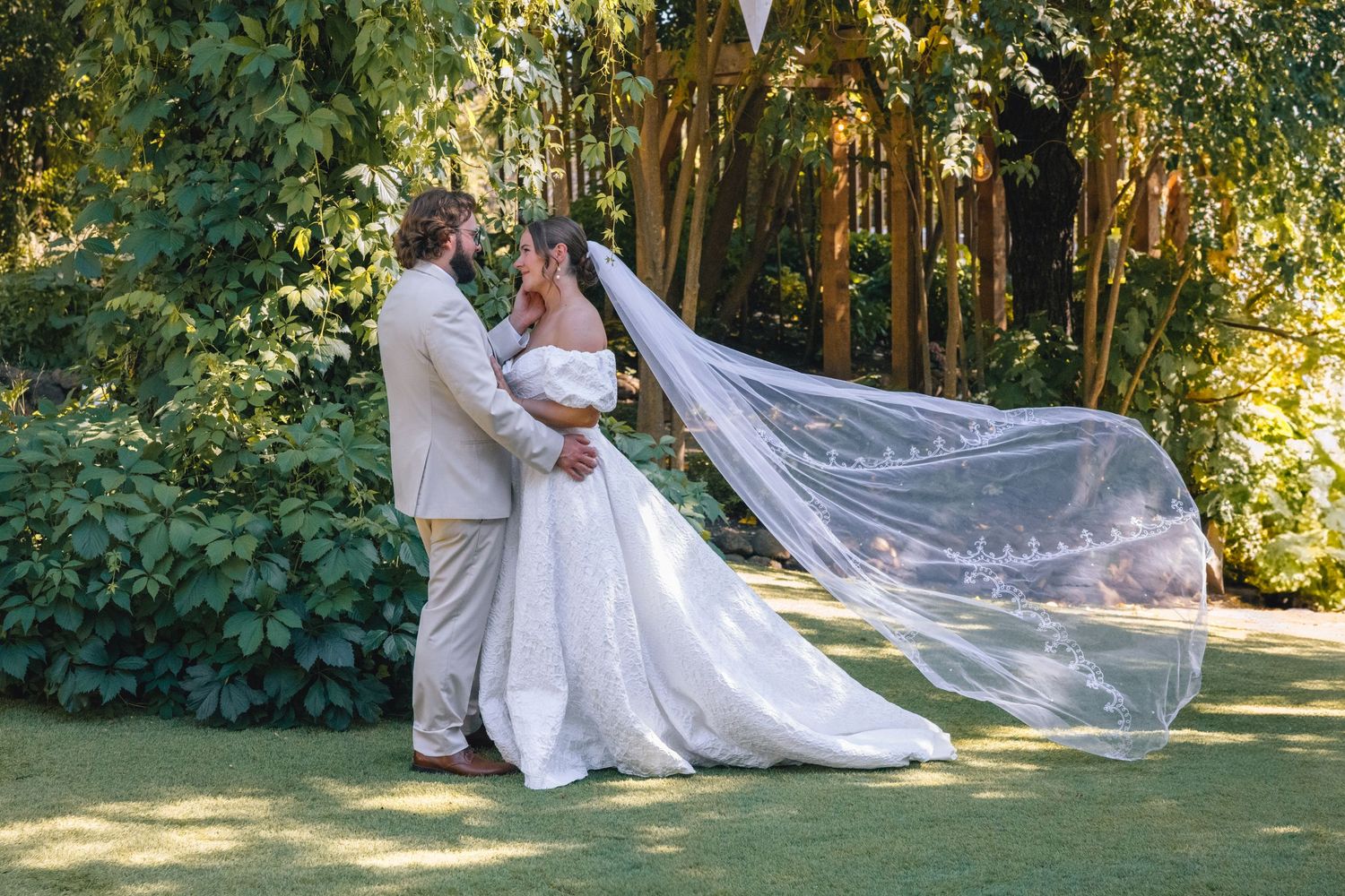 A flowing white wedding dress and veil catch the sunlight in a garden setting.