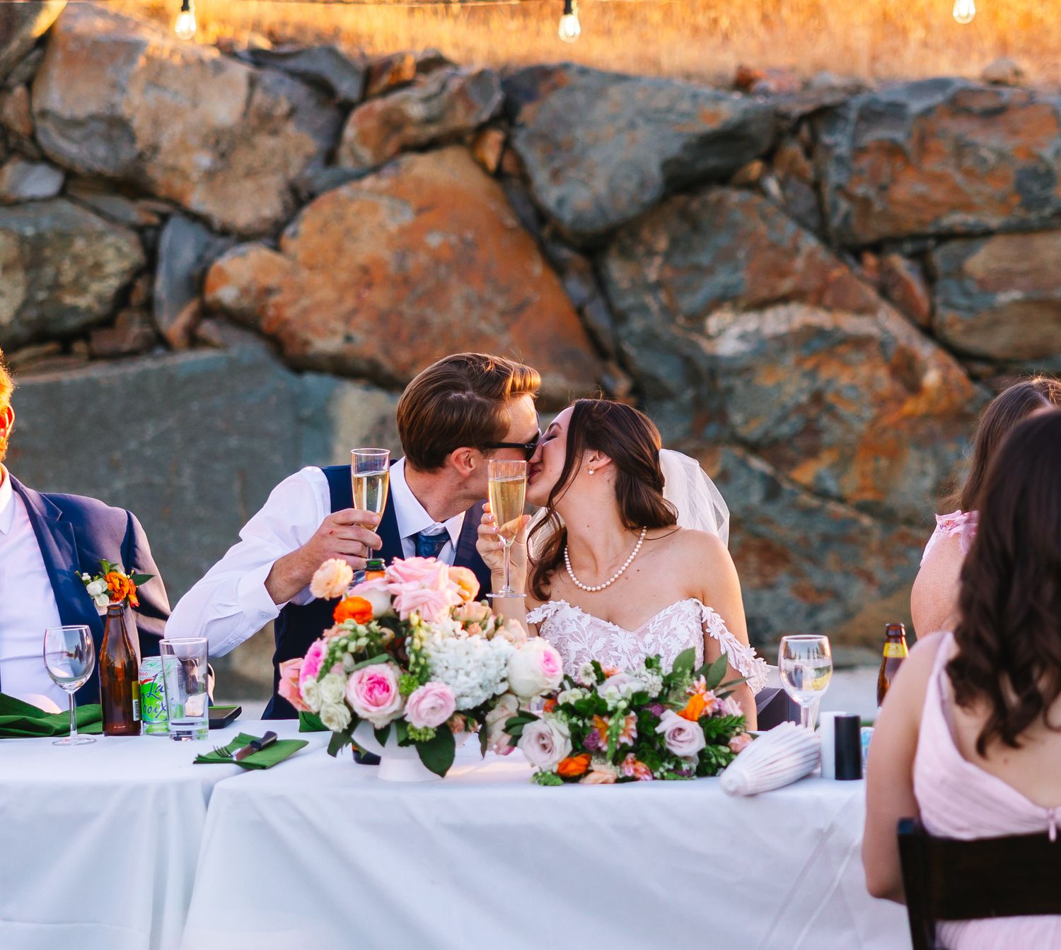 Elegant wedding reception table setup with white linens and floral centerpieces against stone wall backdrop.