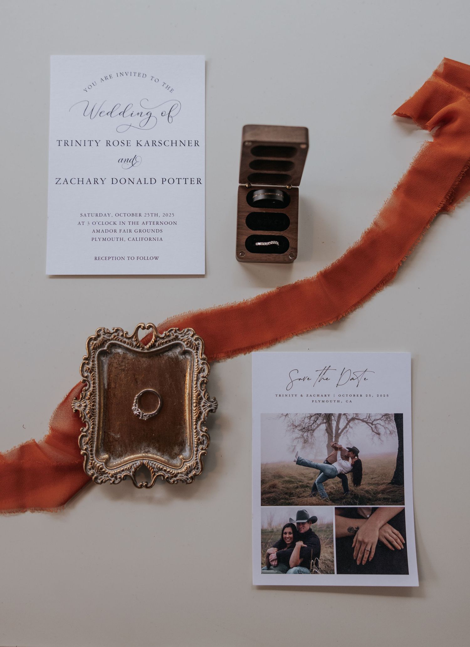 Wedding stationery flatlay with invitation cards, rings, and terracotta silk ribbon on white background.