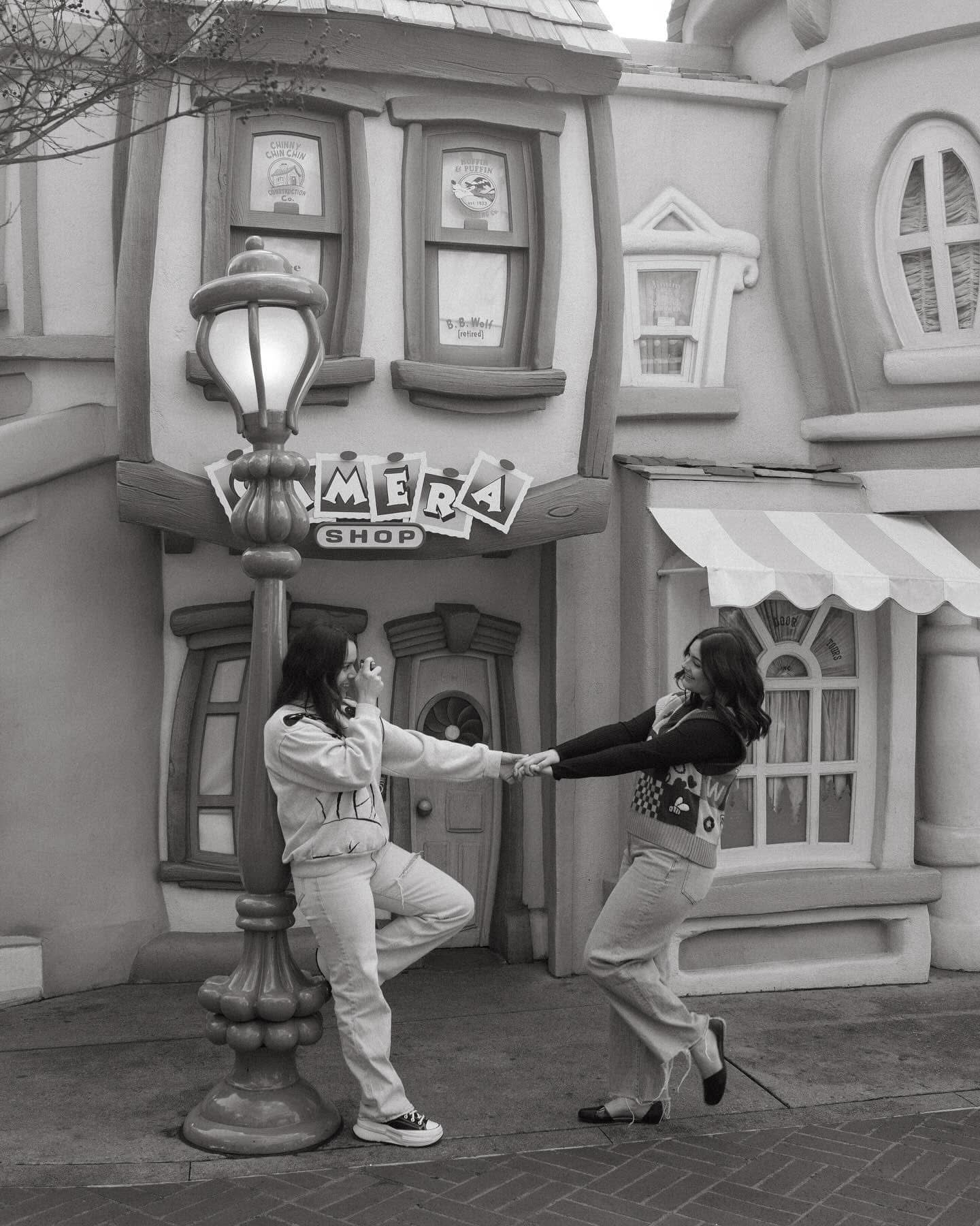 Two women pose playfully near a lamp post outside a camera shop at a theme park in black and white.
