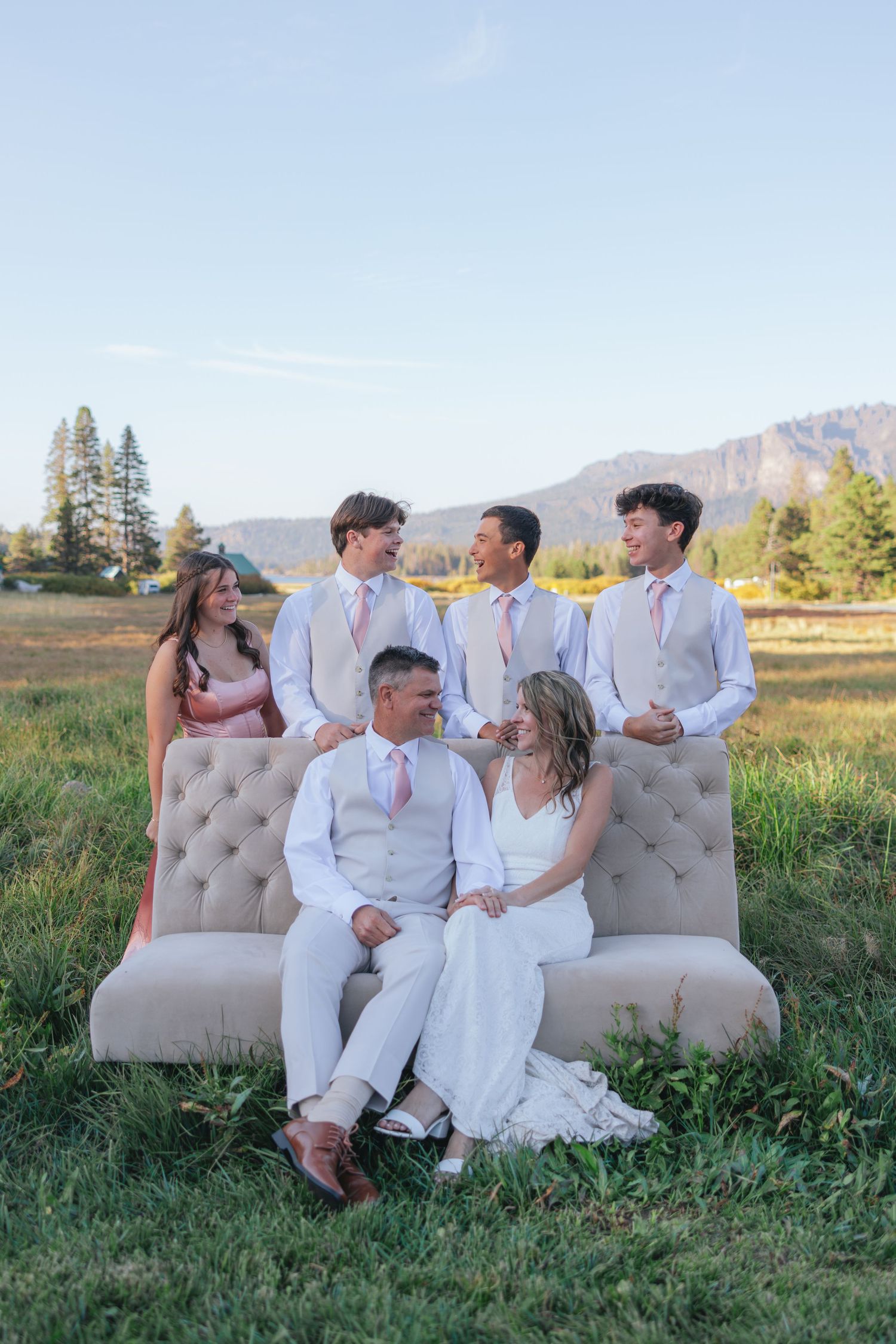 A group in white outfits relaxes on a beige tufted sofa outdoors in a scenic mountain meadow setting at sunset.