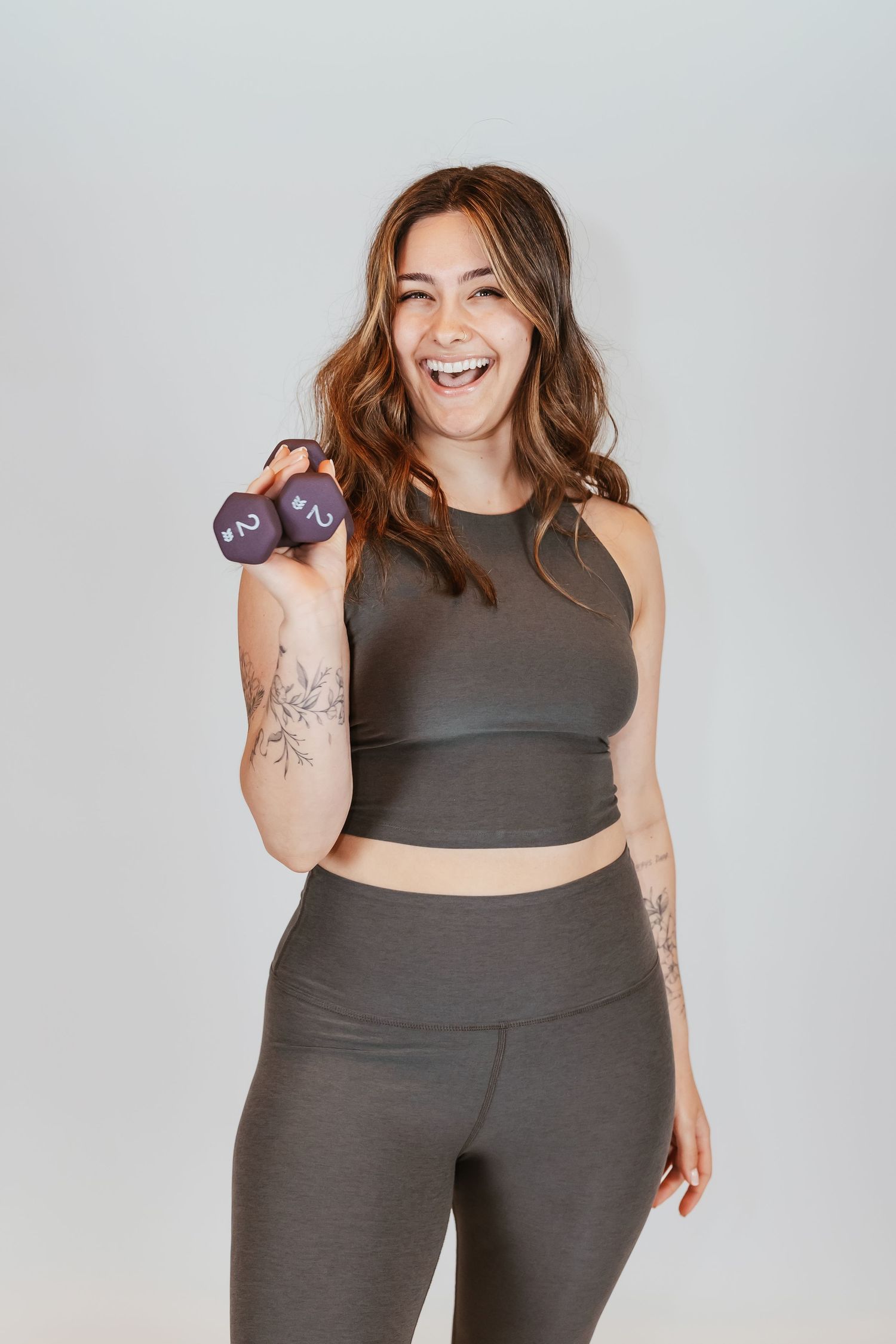 Smiling woman in gray athletic wear holding purple dumbbells, showcasing fitness apparel with visible arm tattoos.