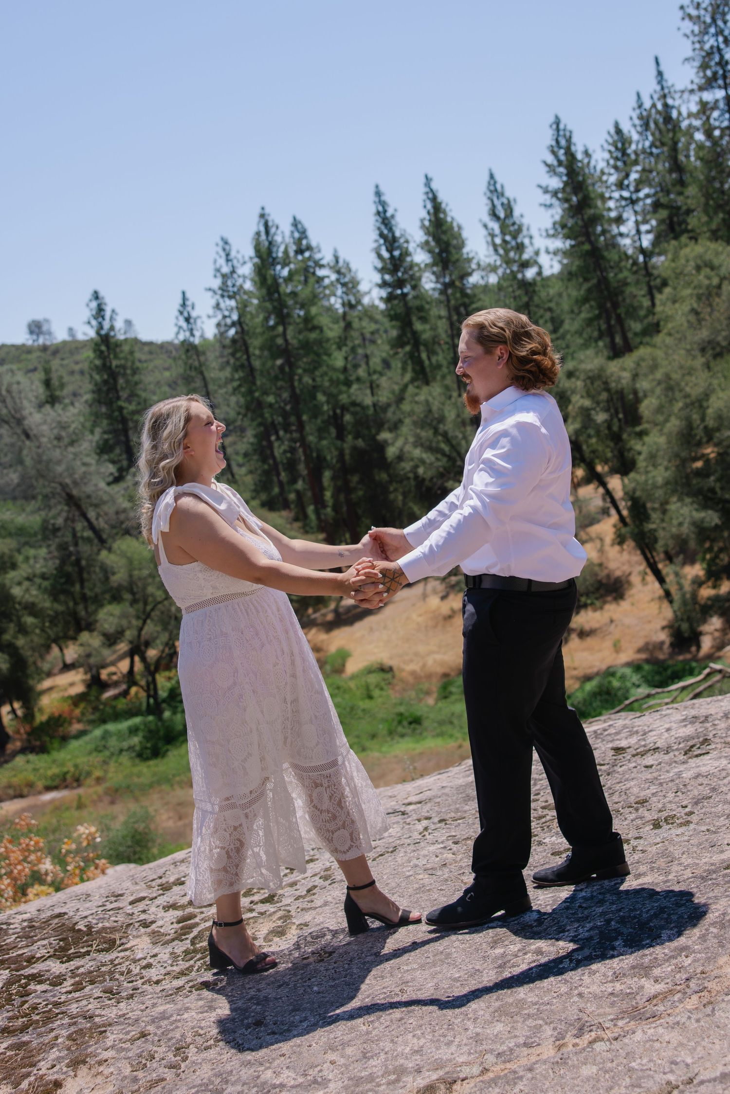 A couple dances together on a rocky outcrop with pine trees in the background.