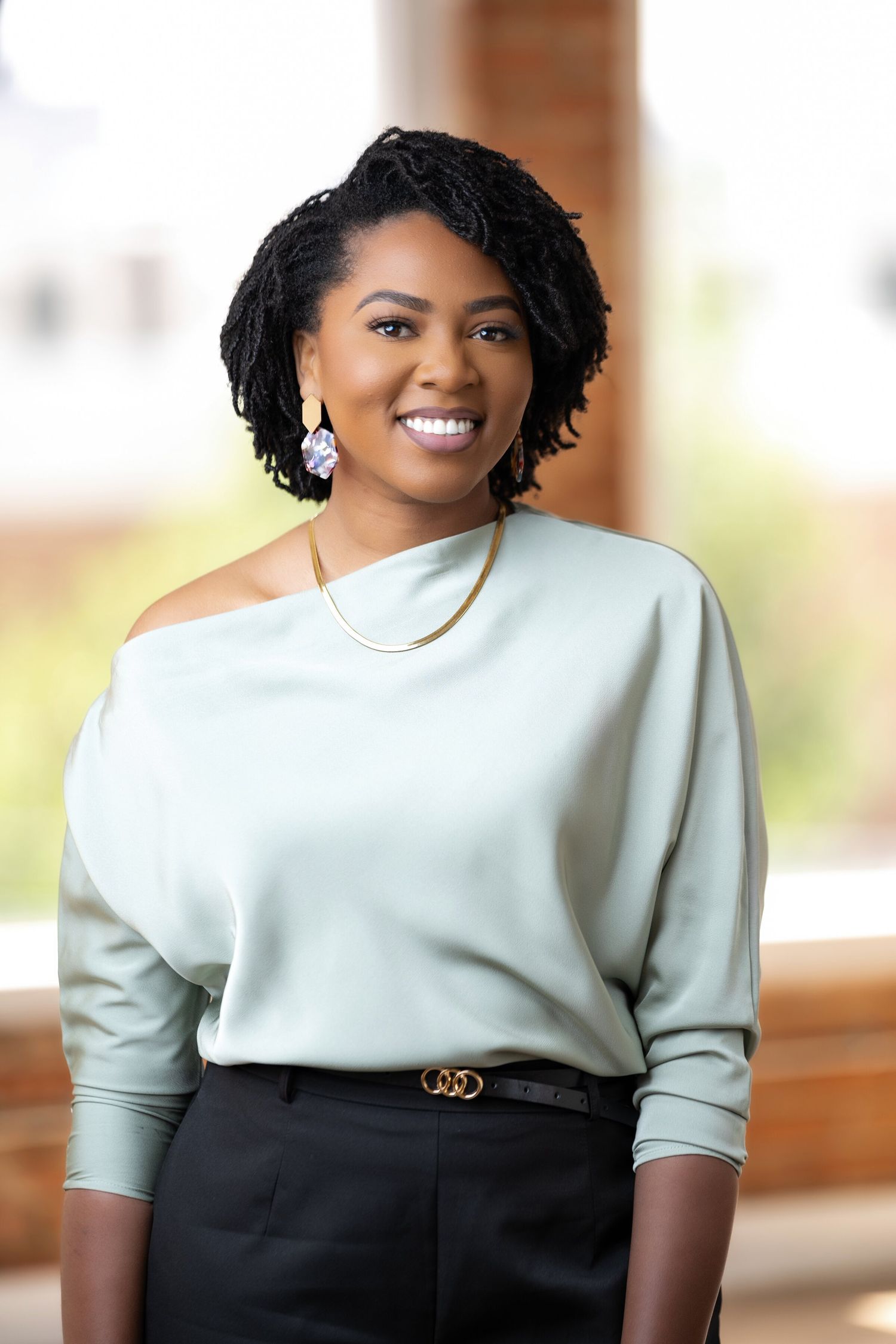 Smiling Black woman with locs wearing a mint off-shoulder top, gold necklace, and black pants in a bright setting.