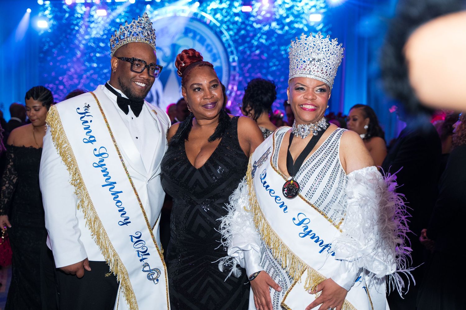 Three formally dressed people pose together at a gala event, two wearing royal crowns and sashes at an elegant celebration.