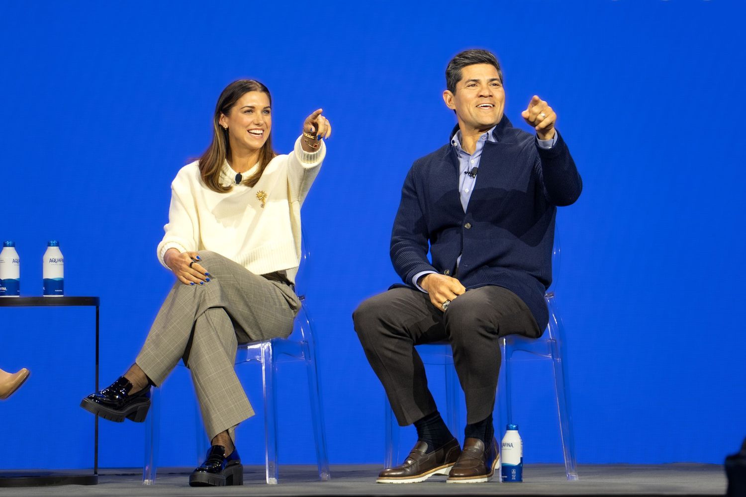 Two people sit on stage pointing forward against a blue background at a corporate event.
