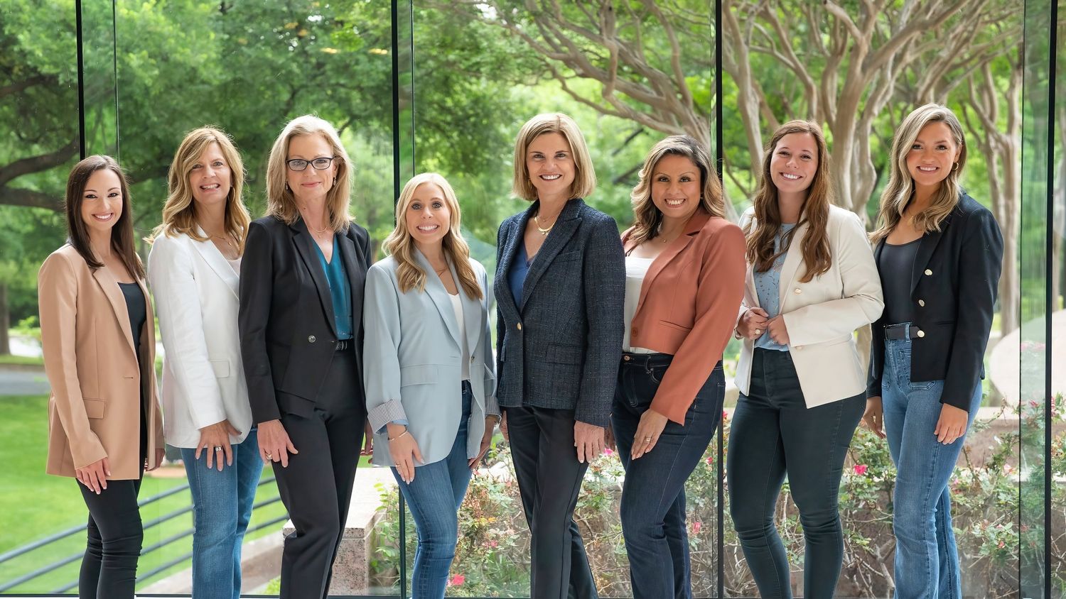 Group of eight professional women posing together outdoors near glass windows in business casual attire.