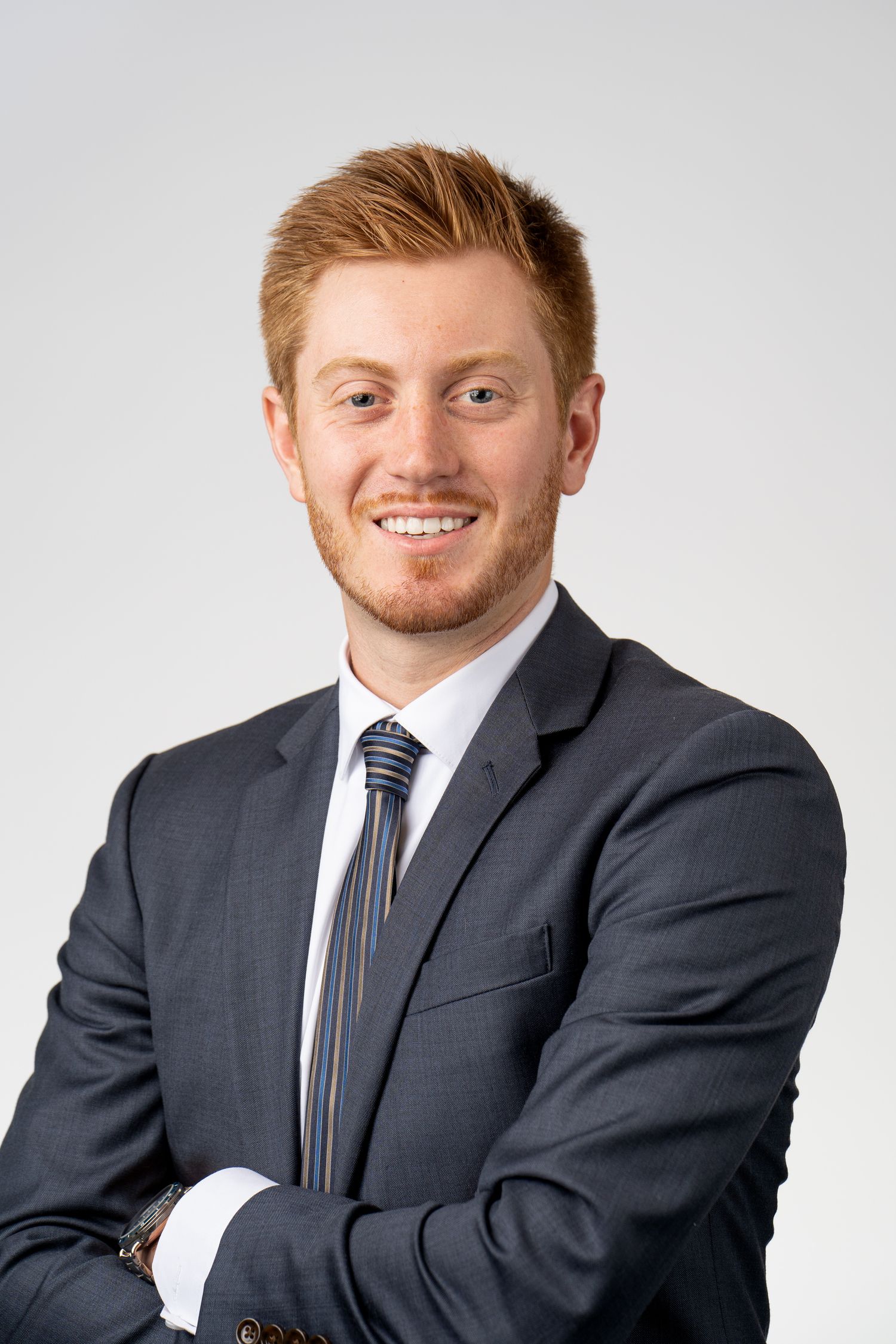 Smiling young professional man with red hair in dark suit and striped tie, arms crossed, against gray background.