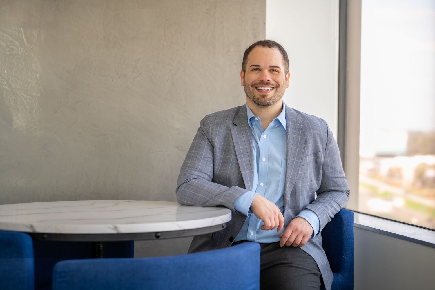 Smiling businessman in gray blazer sitting at marble table by window in modern office setting.
