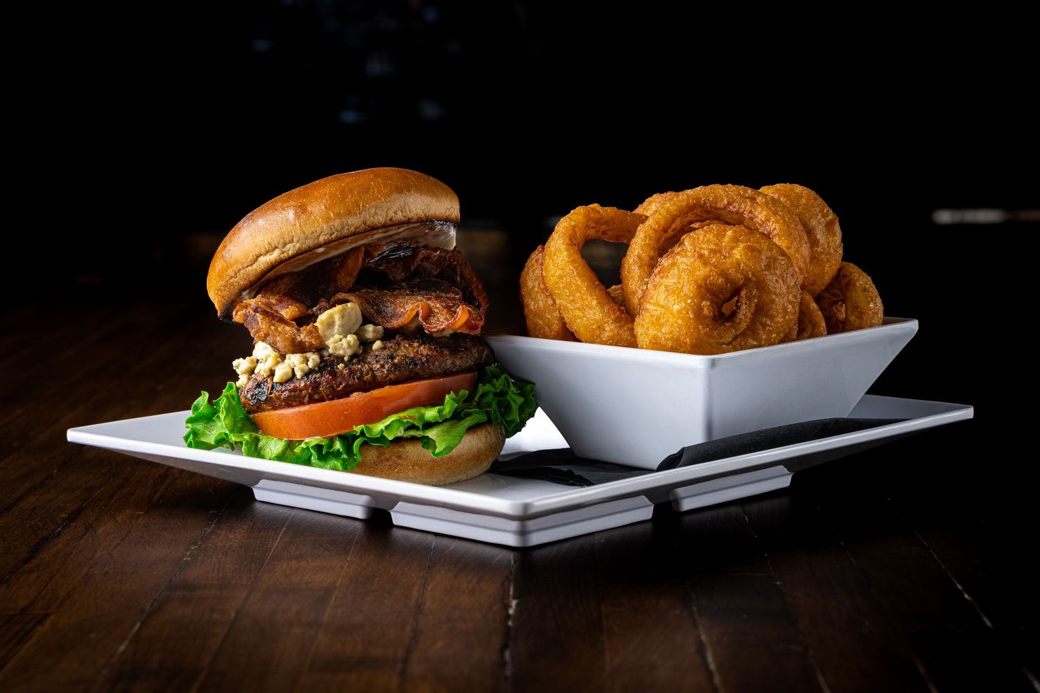 Gourmet burger with toppings on brioche bun beside crispy onion rings in white square bowl on dark wood table.