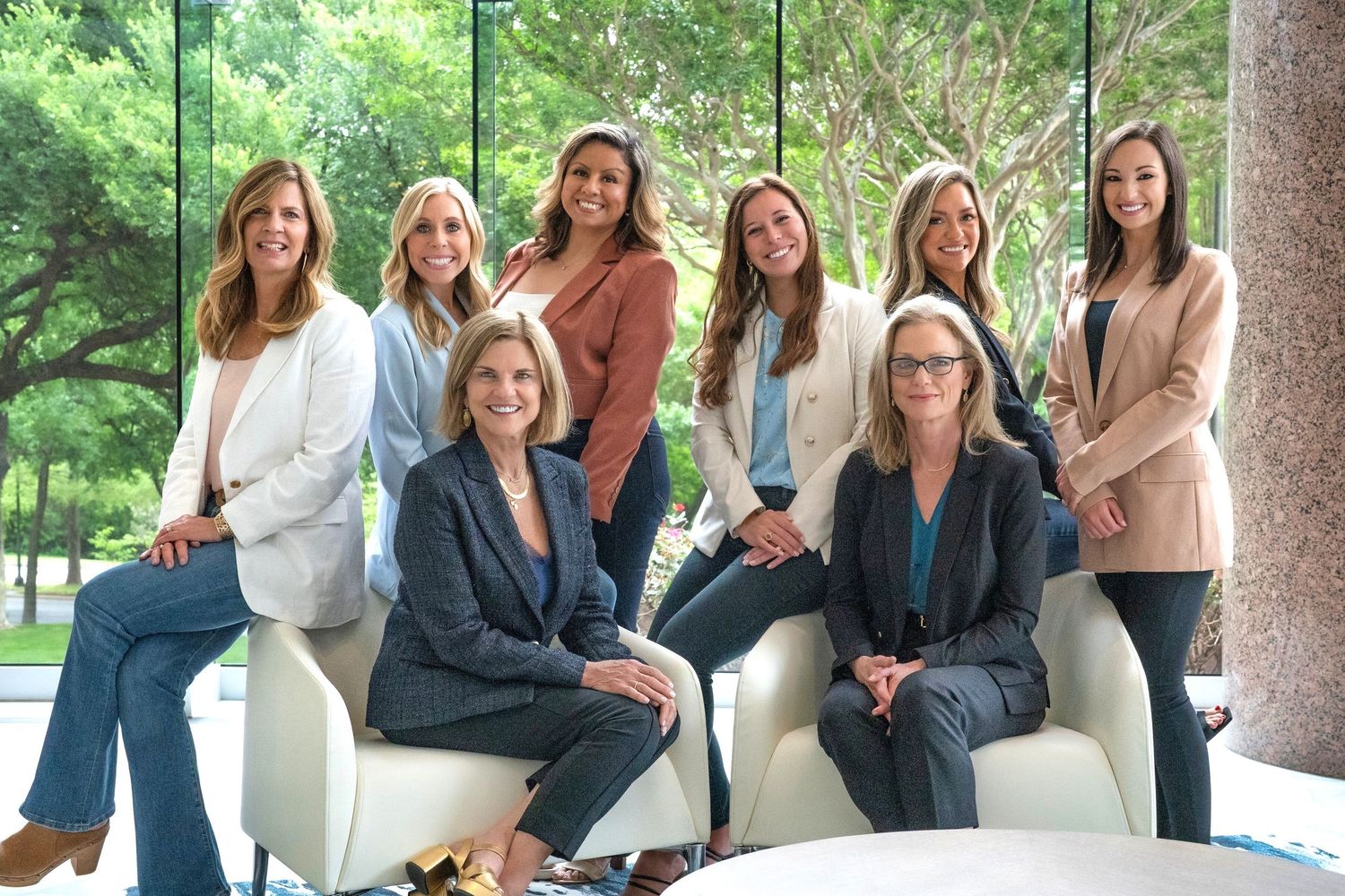 Group of eight professional women posing together in a modern office lobby for a corporate team photo.