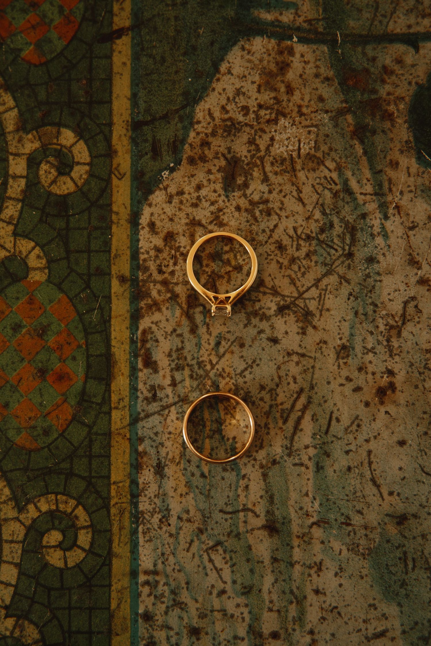 Two gold wedding rings resting against an ornate vintage mosaic background with aged patina details.