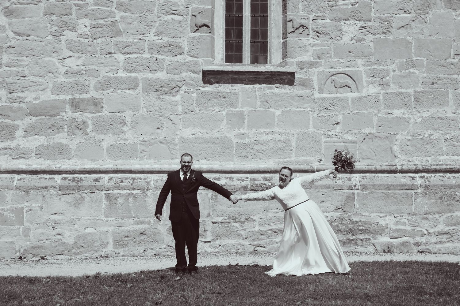 Black and white photo of a bride and groom holding hands outside a stone church on their wedding day.