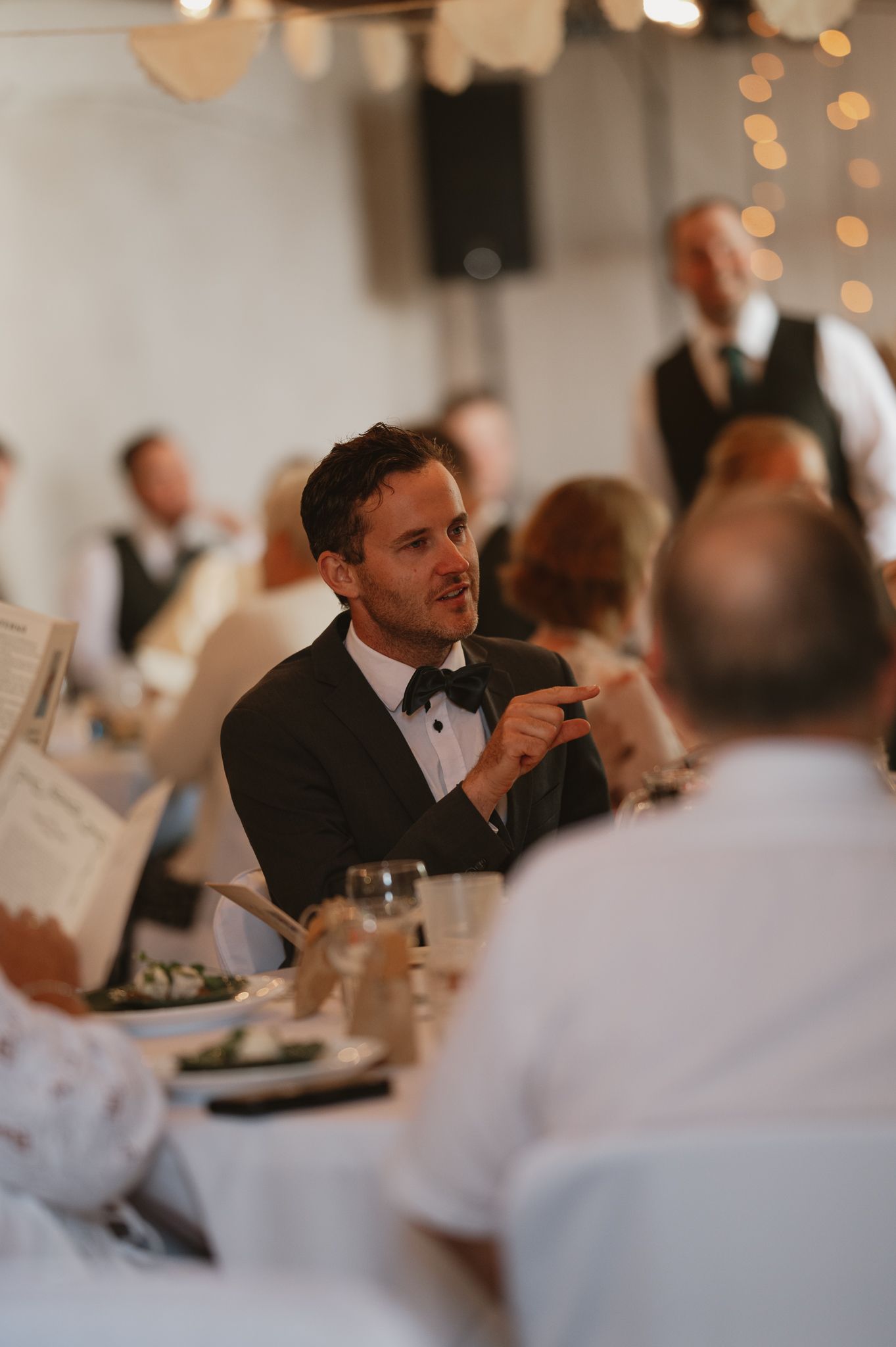 A man in a tuxedo with bow tie gestures while conversing at an elegant wedding reception dinner table.