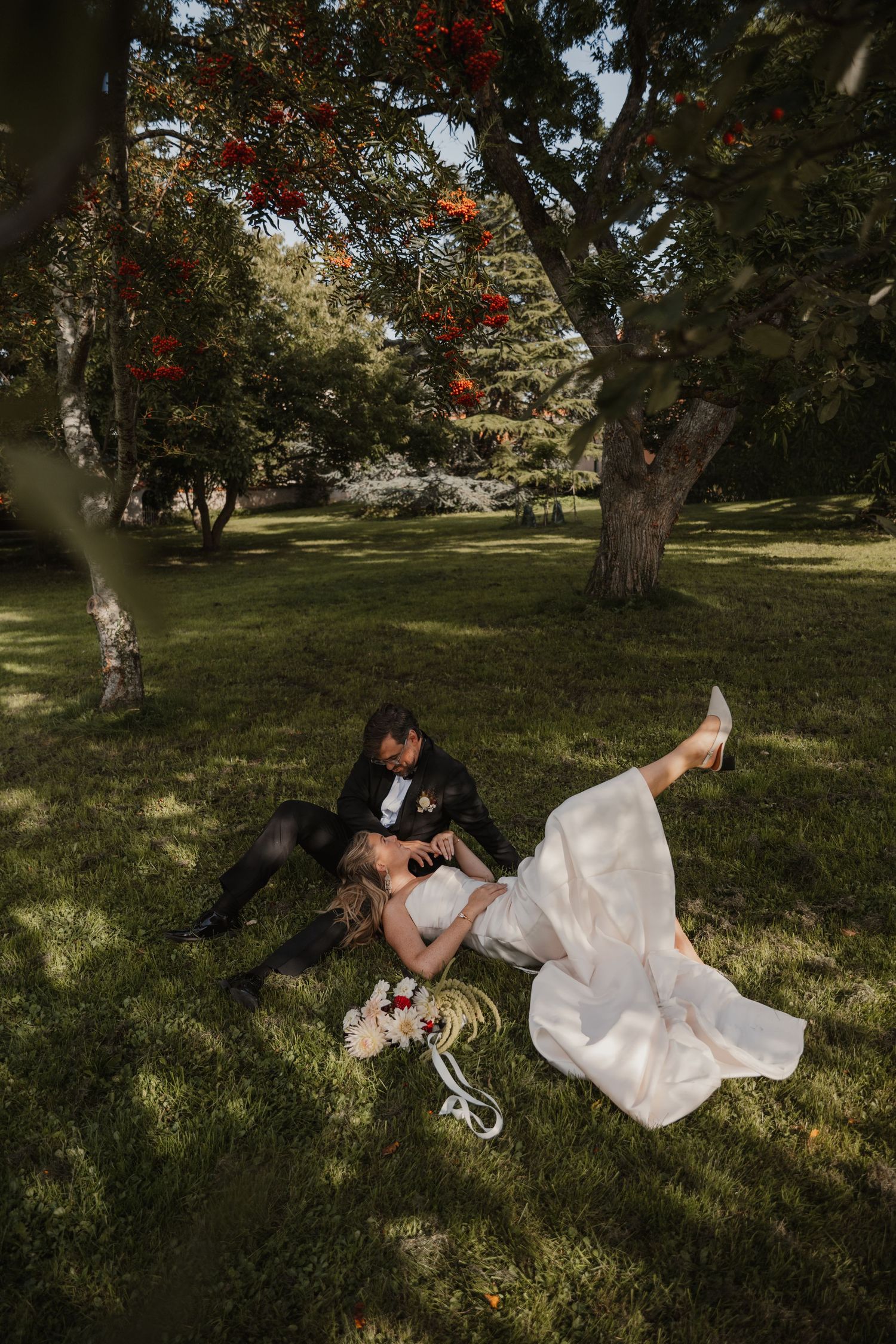 A couple in wedding attire lies together on grassy ground under a tree with red blossoms in a romantic photoshoot.