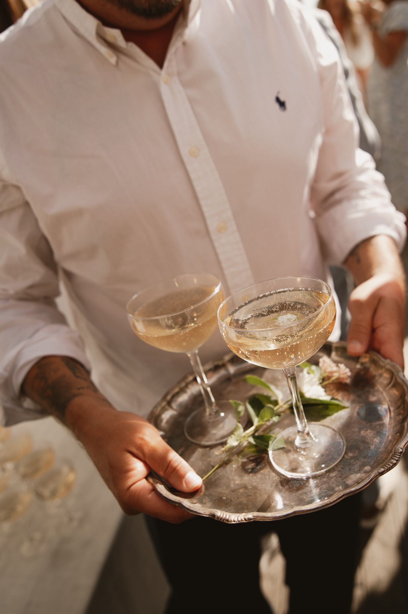 A waiter in a white Ralph Lauren shirt carries a silver tray with two coupe champagne glasses at an elegant event.