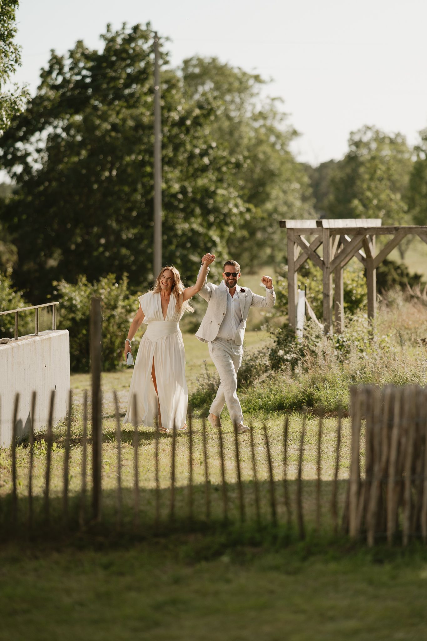 Joyful newlyweds in white attire celebrate outdoors, raising hands on a sunny day in a lush green countryside setting.