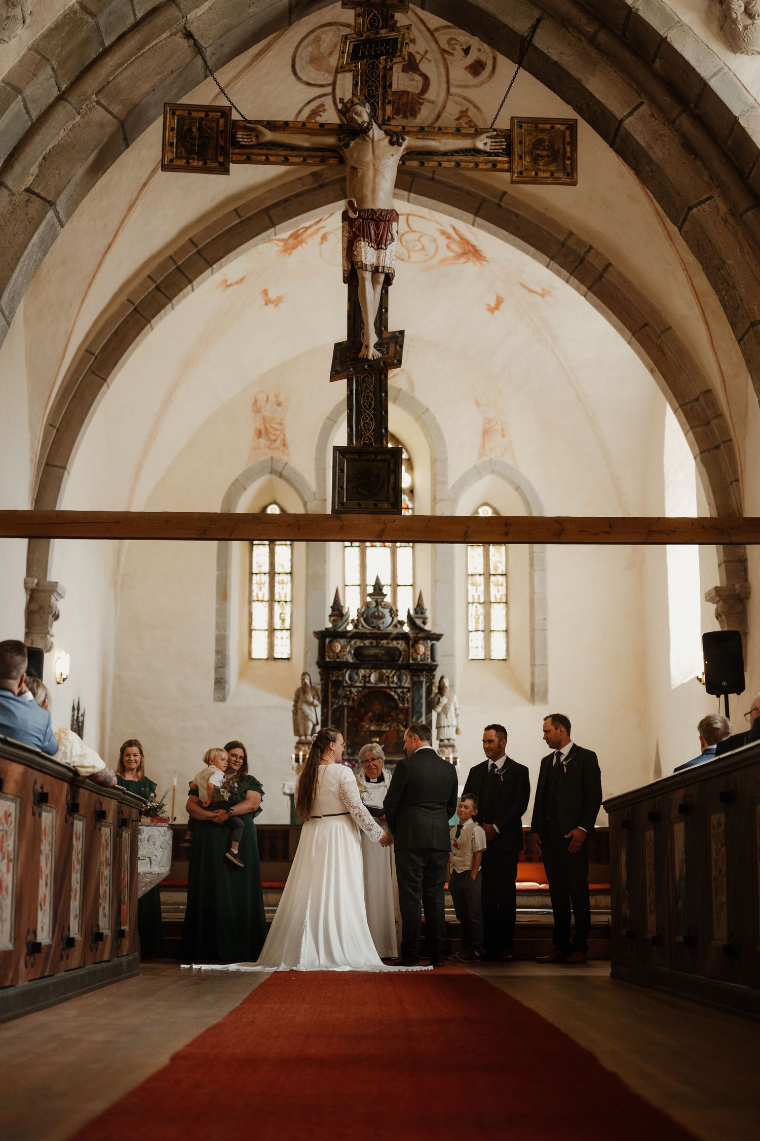 A wedding ceremony in a historic church with a red carpet aisle, bride and groom at the altar beneath a wooden cross.
