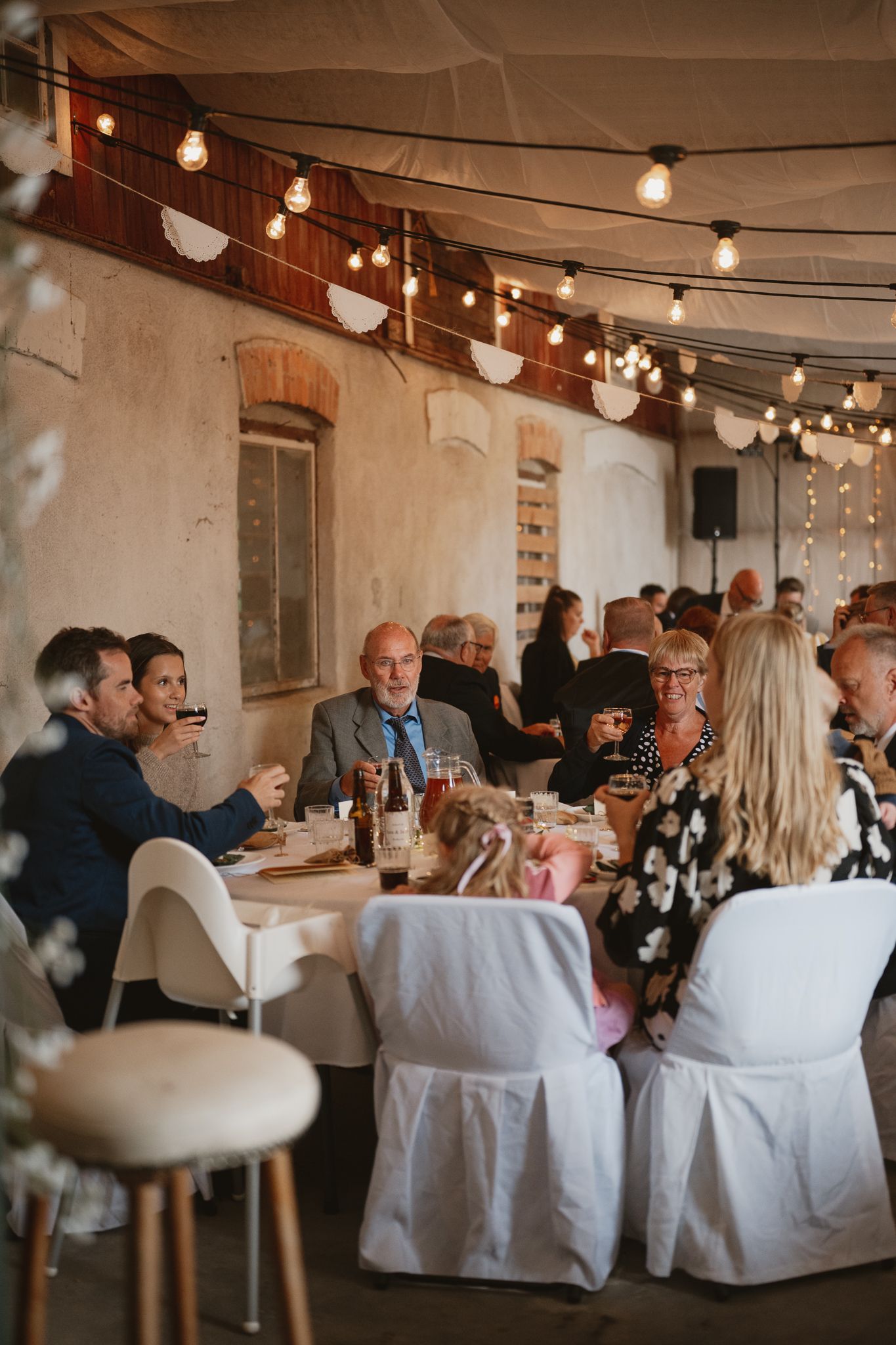 Guests seated at round tables during a wedding reception, decorated with string lights in a rustic venue.