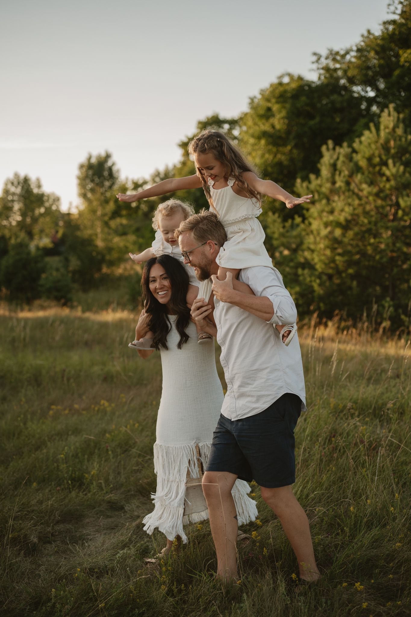 Happy family of four playing outdoors at golden hour, parents carrying children on shoulders in a green meadow.