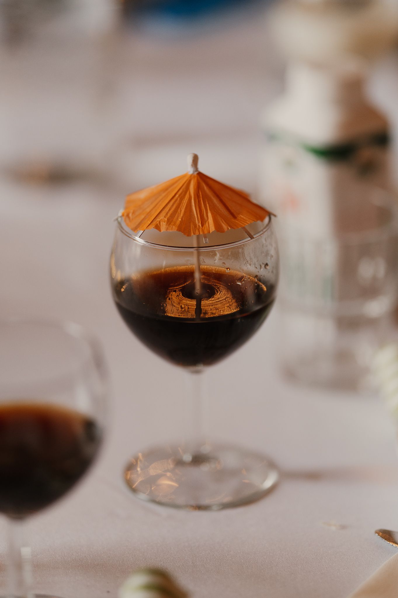 A dark cocktail in a wine glass garnished with a small orange paper umbrella on a white table.
