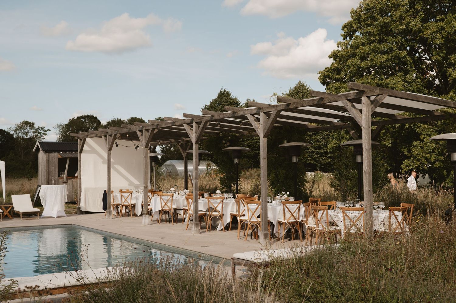 Elegant outdoor wedding reception with white-draped pergola, dining tables, and pool in a lush garden setting.