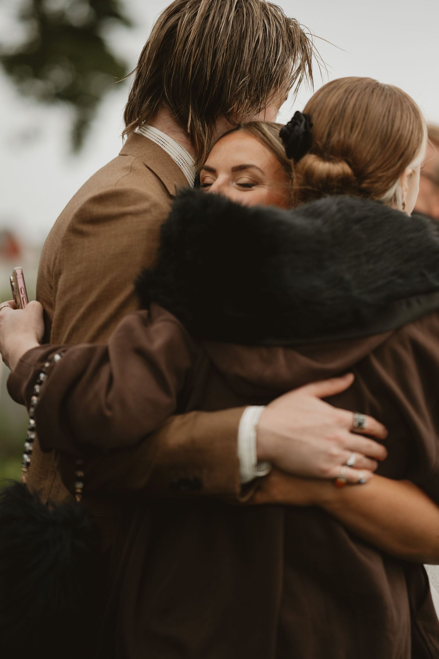 A group of people sharing a warm embrace at an outdoor wedding ceremony, dressed in brown and black coats.
