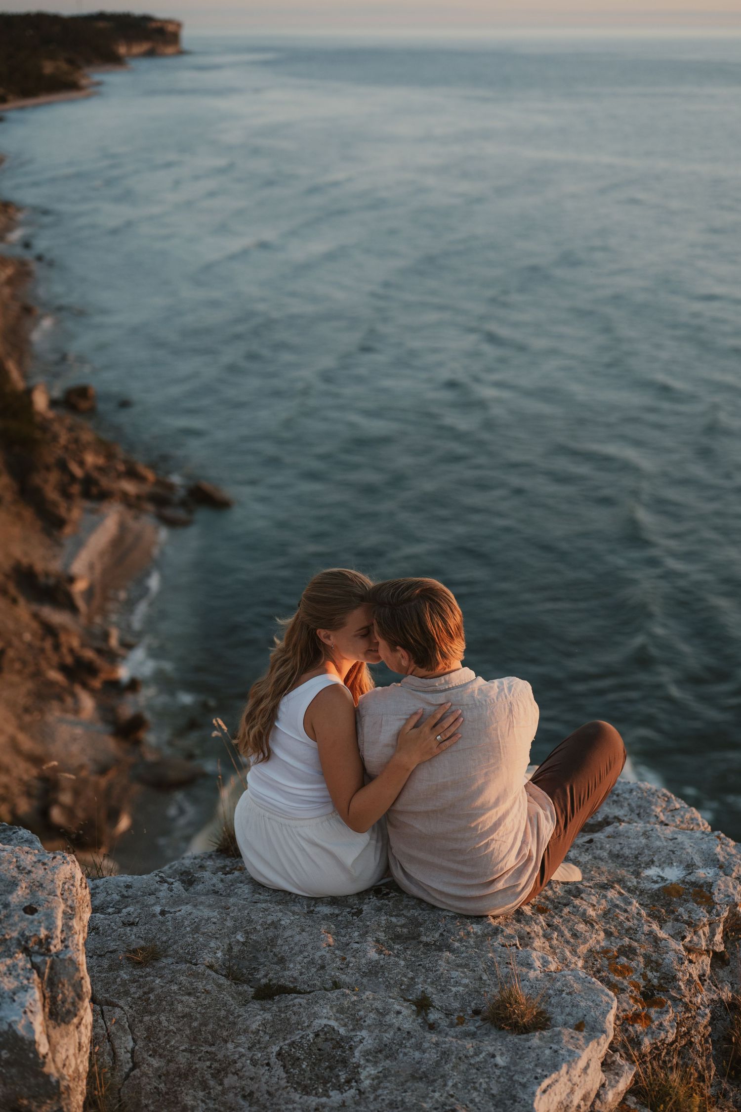 Romantic couple sitting on rocky cliffside overlooking calm ocean at sunset, embracing and sharing a tender moment.